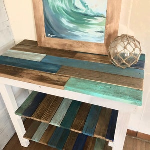 May include: A white wooden table with two shelves. The shelves and top are made of reclaimed wood painted in shades of blue, green, and brown. A framed painting of a wave is on the top shelf, and a glass buoy is on the right side of the table.