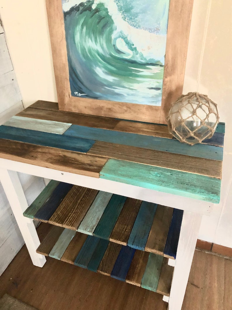 May include: A white wooden table with two shelves. The shelves and top are made of reclaimed wood painted in shades of blue, green, and brown. A framed painting of a wave is on the top shelf, and a glass buoy is on the right side of the table.