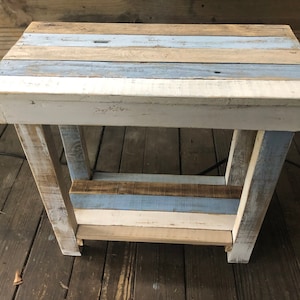 May include: A small, rustic wooden table with a distressed white paint finish. The table top is made of reclaimed wood planks in shades of blue, brown, and white. The table has a single shelf.