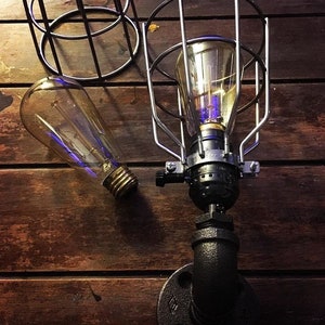 May include: An industrial-style lamp with a clear Edison bulb, encased in a silver metal cage. The lamp is mounted on a black pipe fitting, set against a dark wood background. A second bulb is visible.