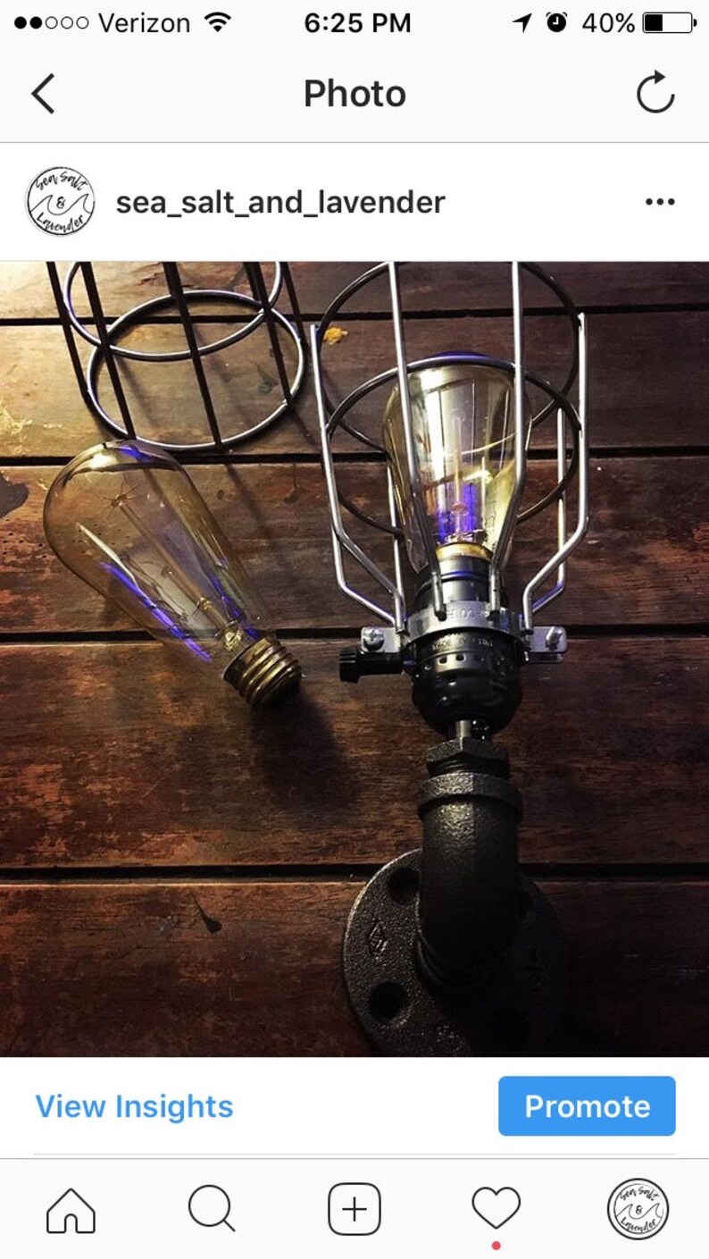 May include: An industrial-style lamp with a clear Edison bulb, encased in a silver metal cage. The lamp is mounted on a black pipe fitting, set against a dark wood background. A second bulb is visible.