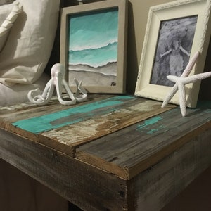May include: A rustic wooden nightstand with a turquoise blue painted top. The nightstand has a white pillow, a white octopus figurine, a framed painting of a beach scene, and a white picture frame with a black and white photo.