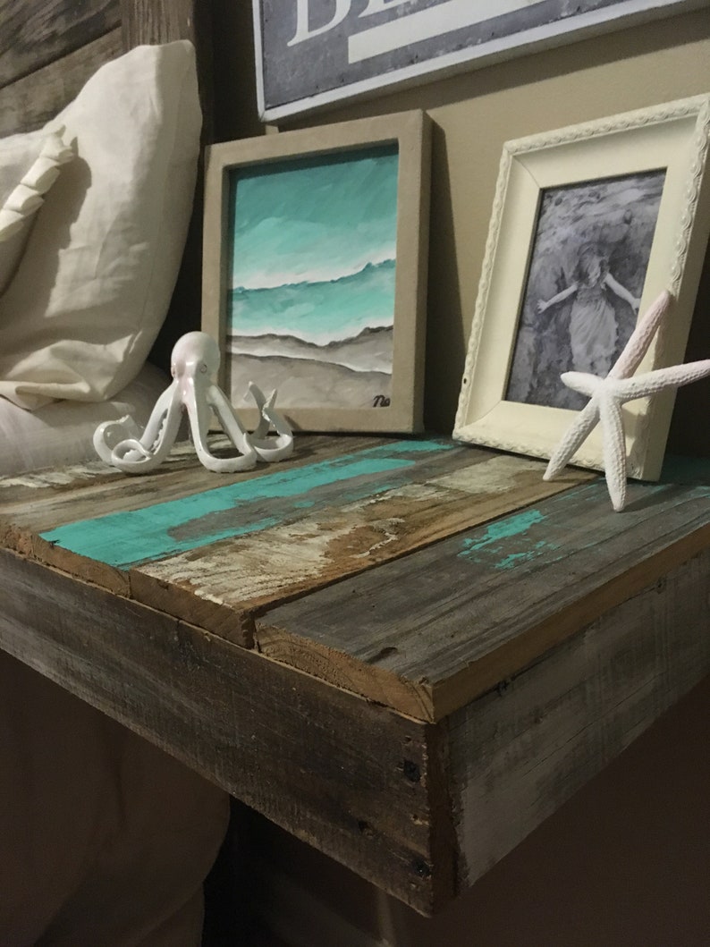 May include: A rustic wooden nightstand with a turquoise blue painted top. The nightstand has a white pillow, a white octopus figurine, a framed painting of a beach scene, and a white picture frame with a black and white photo.