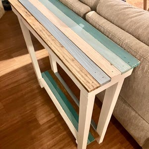 May include: A narrow, rectangular console table with a white frame and a top made of wooden planks in various colors. The planks are painted in shades of blue, teal, and natural wood tones. The table is approximately 48 inches long.