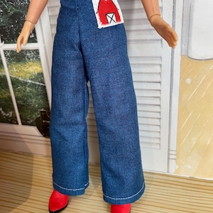 May include: Doll wearing a red and white top, blue denim trousers, and red shoes. The trousers have a patch with a red barn design. The doll's outfit is a playful take on a farm theme.