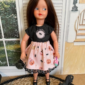 May include: A doll with long brown hair and blue eyes wearing a black short-sleeved top with a circular spider patch and a light pink skirt featuring a pattern of orange pumpkins and black spiders, finished with a black lace trim at the hem.