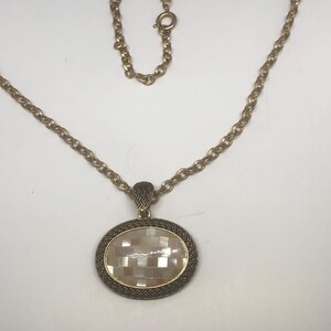 May include: A gold chain necklace with a large oval pendant. The pendant is made of a white shell material with a geometric pattern and a gold metal frame.