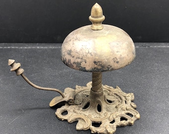 May include: A vintage brass hotel bell with a decorative base and a bell shaped like a dome. The bell has a tarnished finish and a small handle on the side.