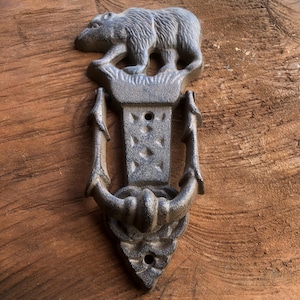 May include: A cast iron door knocker featuring a bear at the top. The door knocker is a dark gray color and has a decorative design. The bear is standing on a decorative base. The knocker is mounted on a wooden door.