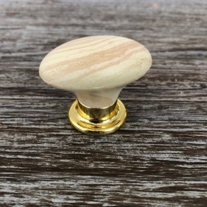 May include: A light brown wooden knob with a gold metal base. The knob has a swirl pattern and is a decorative accent for furniture.