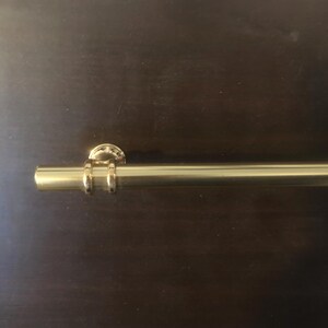 May include: A gold-colored, cylindrical towel bar with a circular wall mount. The bar has a smooth, polished finish and is mounted on a dark brown surface. The bar is designed for hanging towels in a bathroom.
