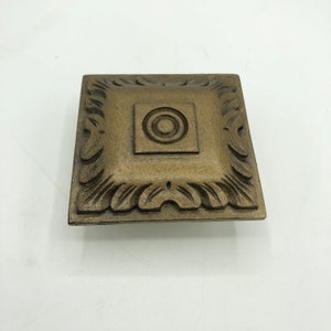 Puede incluir: Una placa cuadrada de metal adornada de color dorado con un diseño circular en relieve en el centro.