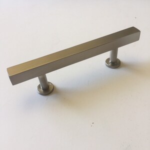 May include: A brushed nickel cabinet pull with a rectangular bar and two round mounting posts. The hardware is a neutral silver color and is designed for use on drawers or cabinet doors. The pull is approximately 6 inches long.