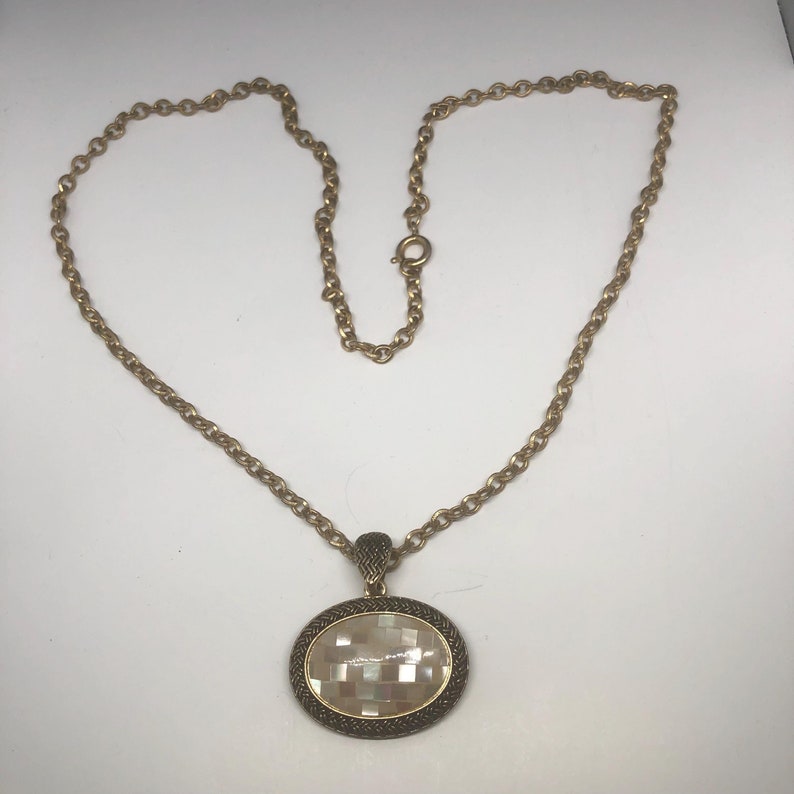 May include: A gold chain necklace with a large oval pendant. The pendant is made of a white shell material with a geometric pattern and a gold metal frame.
