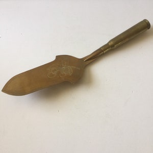 May include: A gold-colored cake server with a cherry design on the blade. The handle is made from a brass bullet casing.