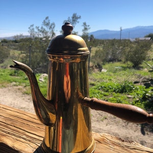 May include: A gold-colored metal coffee pot with a wooden handle. The pot has a rounded, domed lid and a spout that curves upward.