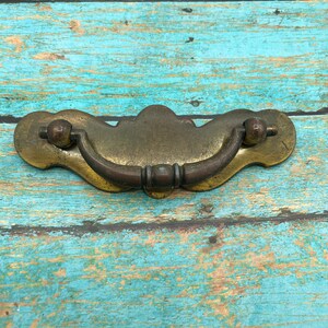 May include: A brass cabinet pull with a curved handle and a decorative design. The pull has a worn, antique finish.