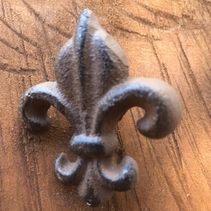 May include: A cast iron fleur-de-lis in a dark brown finish. The decorative emblem is set against a textured, light brown wooden surface. The fleur-de-lis has three petals and curved accents, showcasing a rustic, vintage aesthetic.