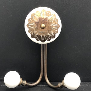 May include: Double hook with a white ceramic base and a gold filigree design. The hook has two white ceramic knobs on the ends.