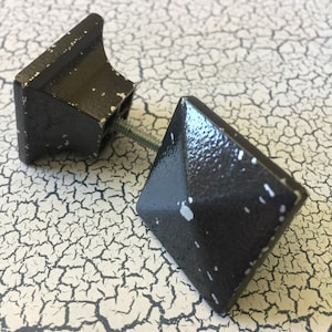 May include: Two black square cabinet knobs with a slightly textured finish. The knobs have a slightly raised center and a screw hole on the back.