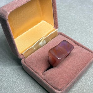 May include: A rectangular, polished gemstone ring with red and purple hues rests in an open, pink velvet ring box. The box has a gold satin interior and is set against a gray surface.
