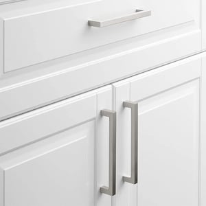 May include: Close-up of white cabinet doors with brushed nickel hardware. The top drawer has a rectangular handle, while the lower doors feature square bar pulls. The hardware provides a modern, minimalist aesthetic.