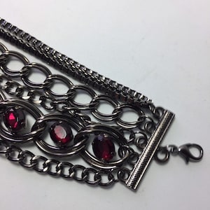 May include: Multi-strand bracelet with a black metal chain and red gemstone accents. The bracelet has a toggle clasp.