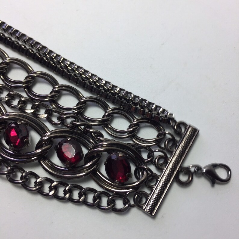 May include: Multi-strand bracelet with a black metal chain and red gemstone accents. The bracelet has a toggle clasp.