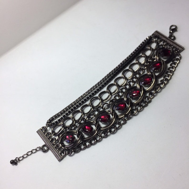 May include: A multi-layered chain bracelet with a dark gunmetal finish and red gemstone accents.