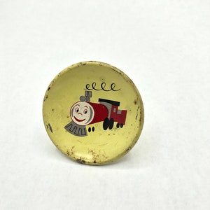 May include: A yellow metal trinket with a cartoon train design. The train is red with a smiling face and black wheels. The train is on a yellow background with the word "eee" above it.