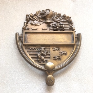 Op de afbeelding: Een bronzen deurklopper met een schildontwerp. De klopper heeft een ridderhelm, decoratief krulwerk en een rechthoekige ruimte voor een naam of inscriptie. De onderkant heeft een stervormig ontwerp.