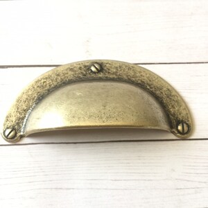May include: A gold-toned metal cabinet pull with a rounded, slightly domed shape. The pull has a textured surface and four visible screws.