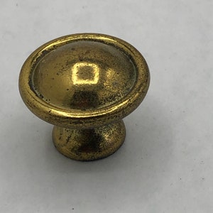 May include: A gold-toned metal knob with a round, slightly domed top and a raised square in the center. The knob has a smooth, slightly textured surface.