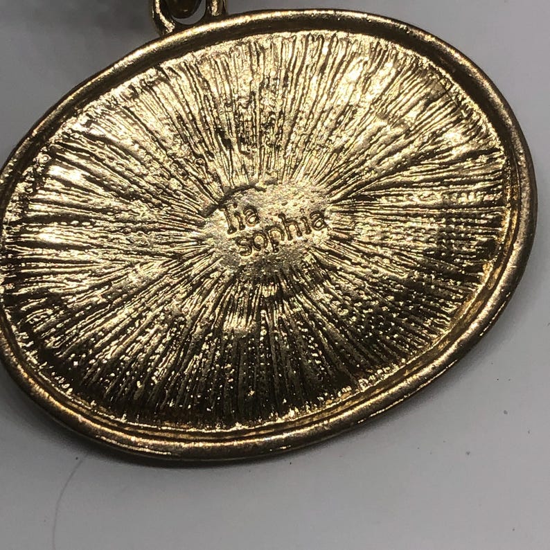 May include: Gold-toned oval pendant with a textured surface and the inscription "Tia Sophia" on the back.