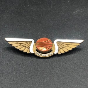 May include: A gold and silver winged pin with a red and orange globe in the center. The globe has a white stripe across it. The words "CONTINENTAL" are written in a white arc below the globe.