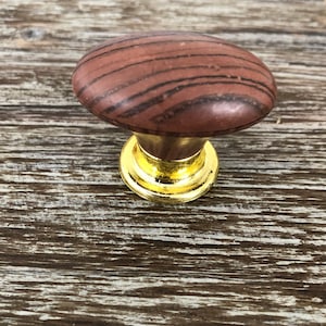 May include: An oval-shaped wooden cabinet knob with a brown wood grain pattern and a gold metal base.