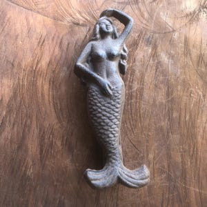 May include: A cast iron mermaid figurine with a textured, weathered appearance. The mermaid has long flowing hair, a scaled tail, and is posed with one arm raised. The figurine is set against a wooden background.