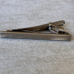 May include: A silver-toned metal tie clip featuring a sleek, elongated rectangular bar design with two horizontal engraved lines running along its length. The accessory includes a spring-loaded hinge mechanism on the back for secure attachment.