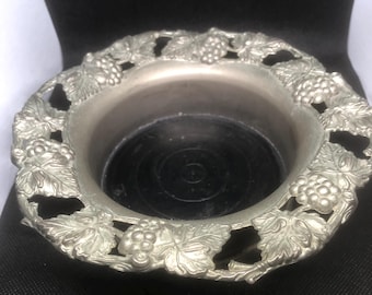 May include: Silver-toned metal wine bottle coaster with a grapevine design. The coaster has a raised rim and a flat bottom.