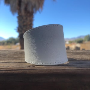 May include: White leather cuff bracelet with a stitched edge. The bracelet is curved and has a textured surface.