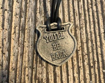May include: A silver-toned pendant necklace with a shield-shaped charm that reads "You'll Be Back". The pendant is attached to a black leather cord.