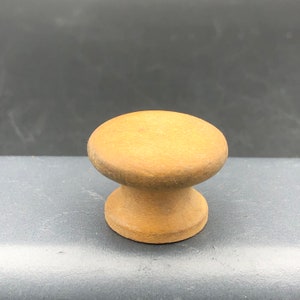 May include: A wooden knob with a rounded top and a slightly flared base. The knob is a light brown color and has a smooth, polished finish.
