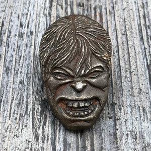 May include: A silver-colored metal sculpture of a face with a furrowed brow and a wide, toothy grin. The sculpture is detailed with hair and facial features.