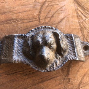 May include: A cast iron dog head door pull with a weathered, textured finish. The dog's face is detailed, with visible fur and a slightly open mouth. The pull has a rectangular base with a hole for mounting.