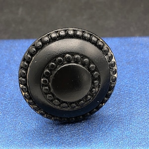 May include: Black round cabinet knob with a raised, concentric circle design. The knob has a smooth, matte finish.