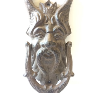 Può includere: Un battente per porta in ghisa a forma di volto grottesco con corna e baffi. Il battente è di colore marrone scuro e usurato e ha una maniglia a forma di anello sotto il volto.