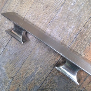 May include: A silver metal door handle with two rectangular pulls. The handle is long and narrow, with a brushed finish.