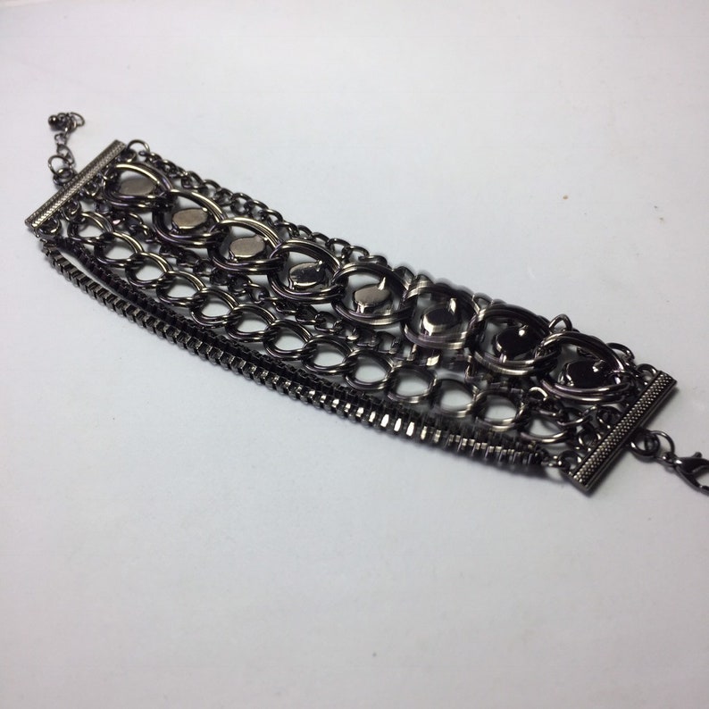 May include: A multi-layered bracelet with a black metal finish. The bracelet features a variety of chain styles and decorative elements, including heart-shaped charms.