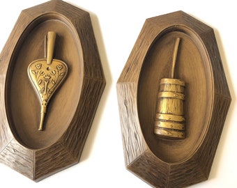 May include: A pair of wall decor pieces in a woodgrain finish. Each piece features a raised oval with a gold-colored object inside. The left piece features a bellows and the right piece features a bucket.