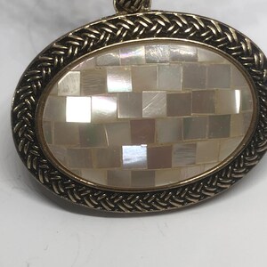 May include: A gold oval pendant with a braided border and a center made of white mother of pearl tiles.